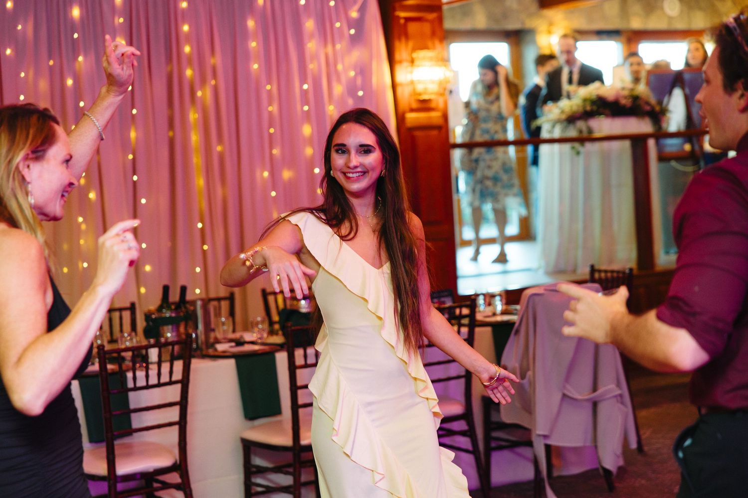 Bride in white dress dancing joyfully with guests at elegant wedding reception with string lights.
