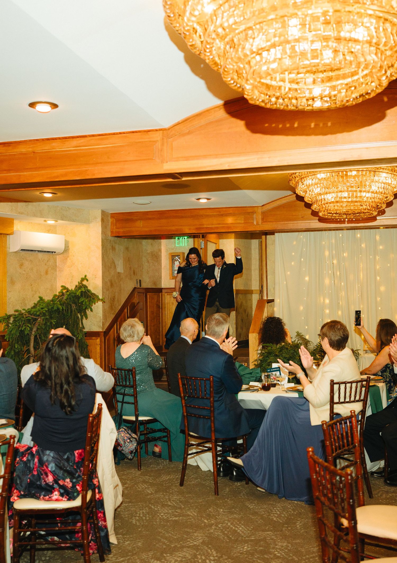 Wedding guests applaud as a couple makes their grand entrance down the stairs at a warmly lit reception hall.