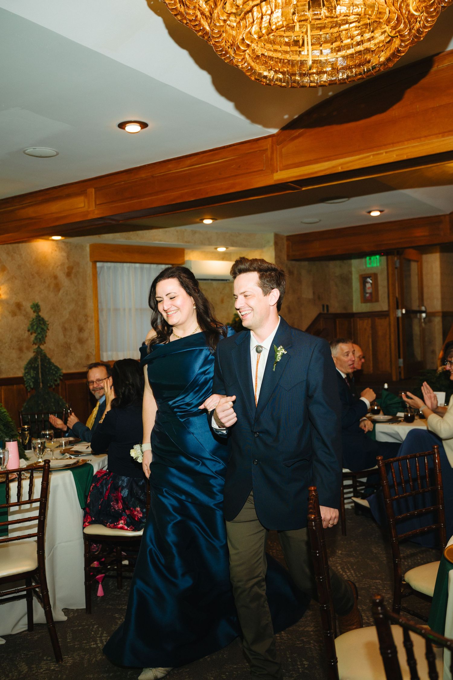 Couple walks arm-in-arm through elegant restaurant reception hall with chandelier lighting during wedding celebration.
