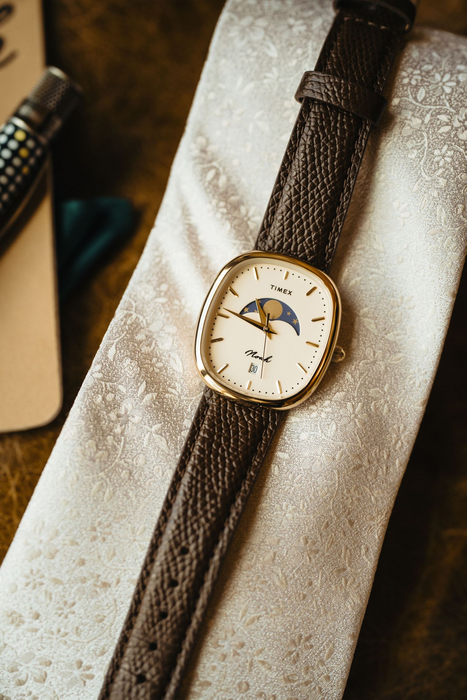 Vintage Timex gold moon phase watch with brown leather strap resting on white floral fabric.