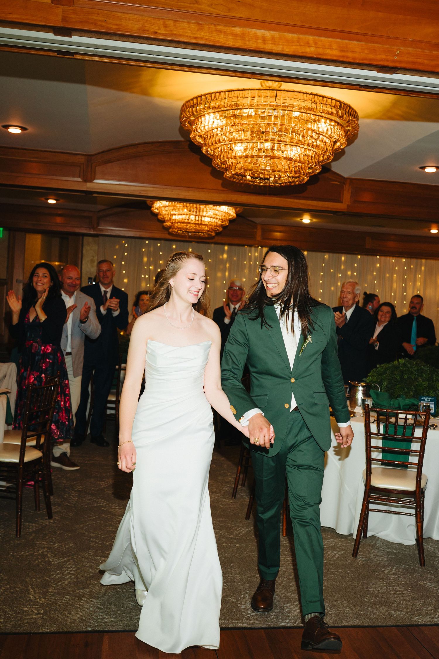 Bride in white gown and groom in green suit hold hands entering wedding reception under chandelier.