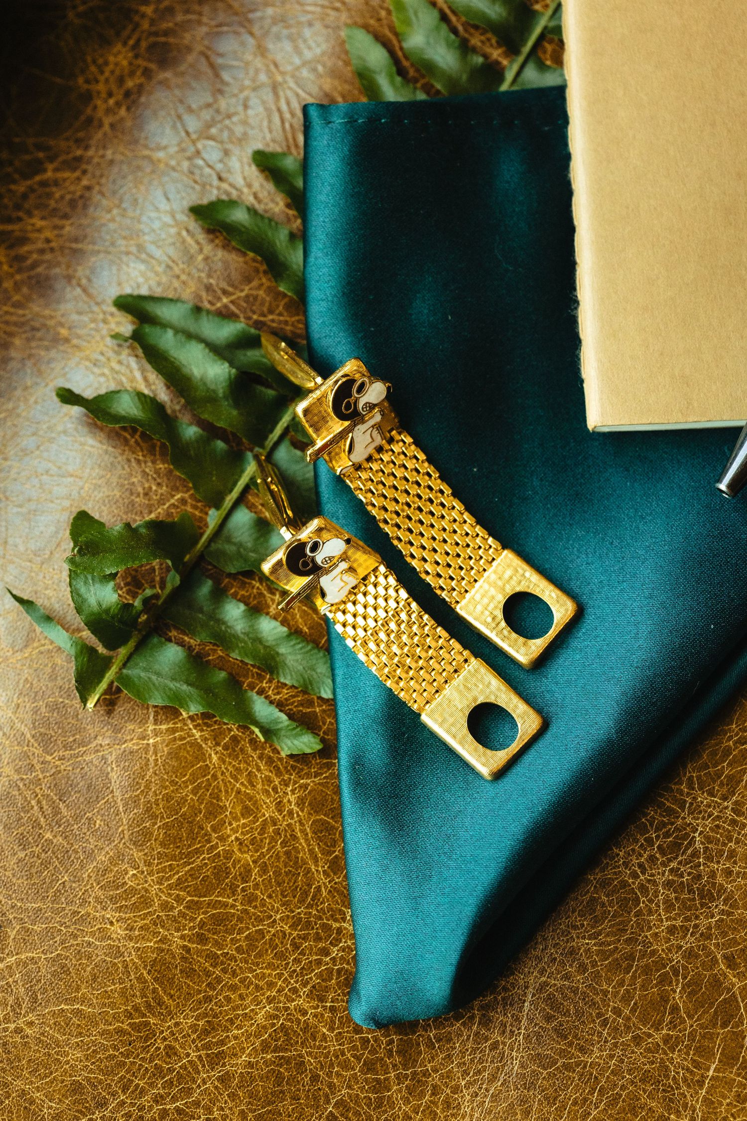 Gold mesh suspenders and teal pocket square with green fern leaves on brown leather background.