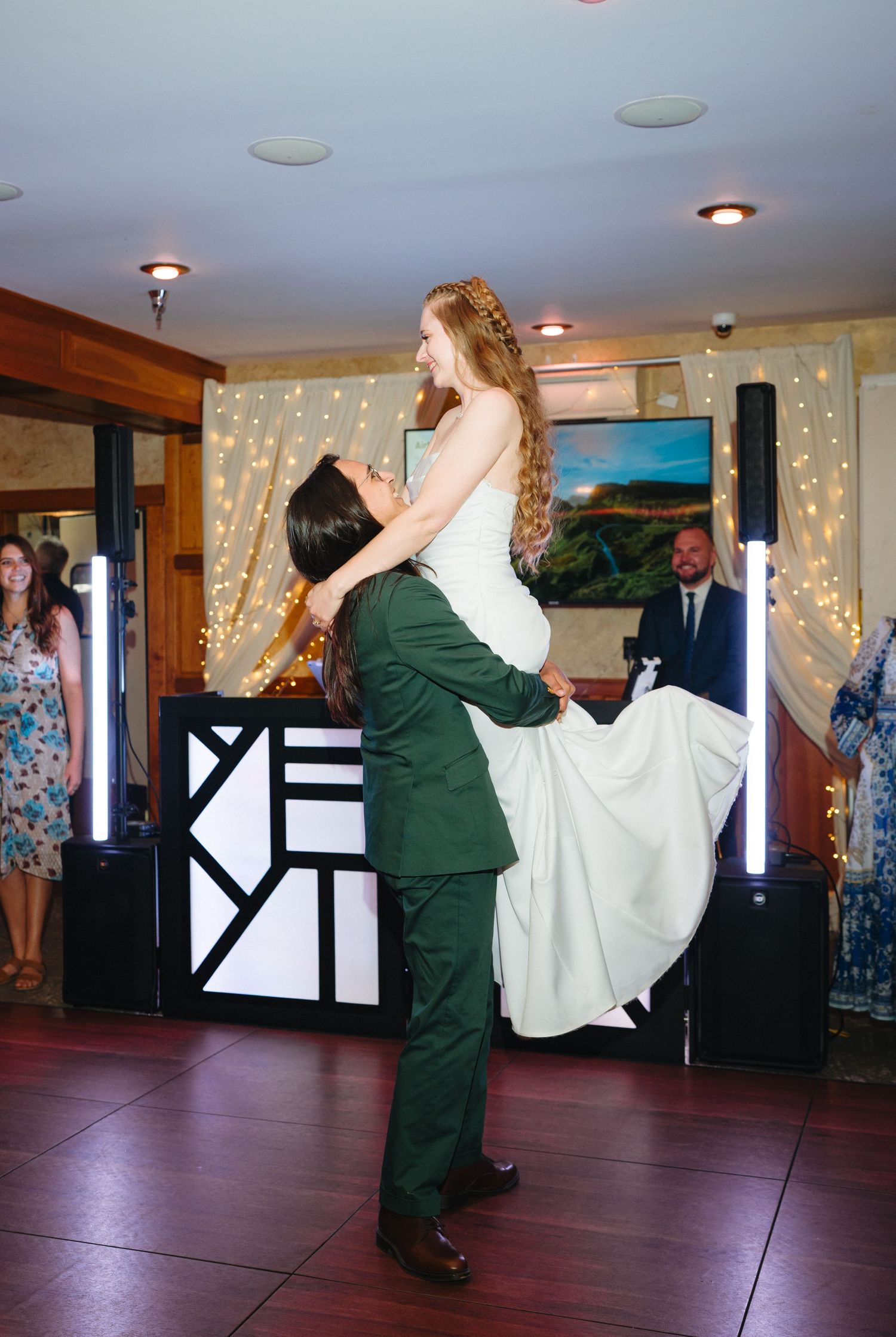 Groom in green suit lifts bride in white wedding dress during first dance at elegant reception.