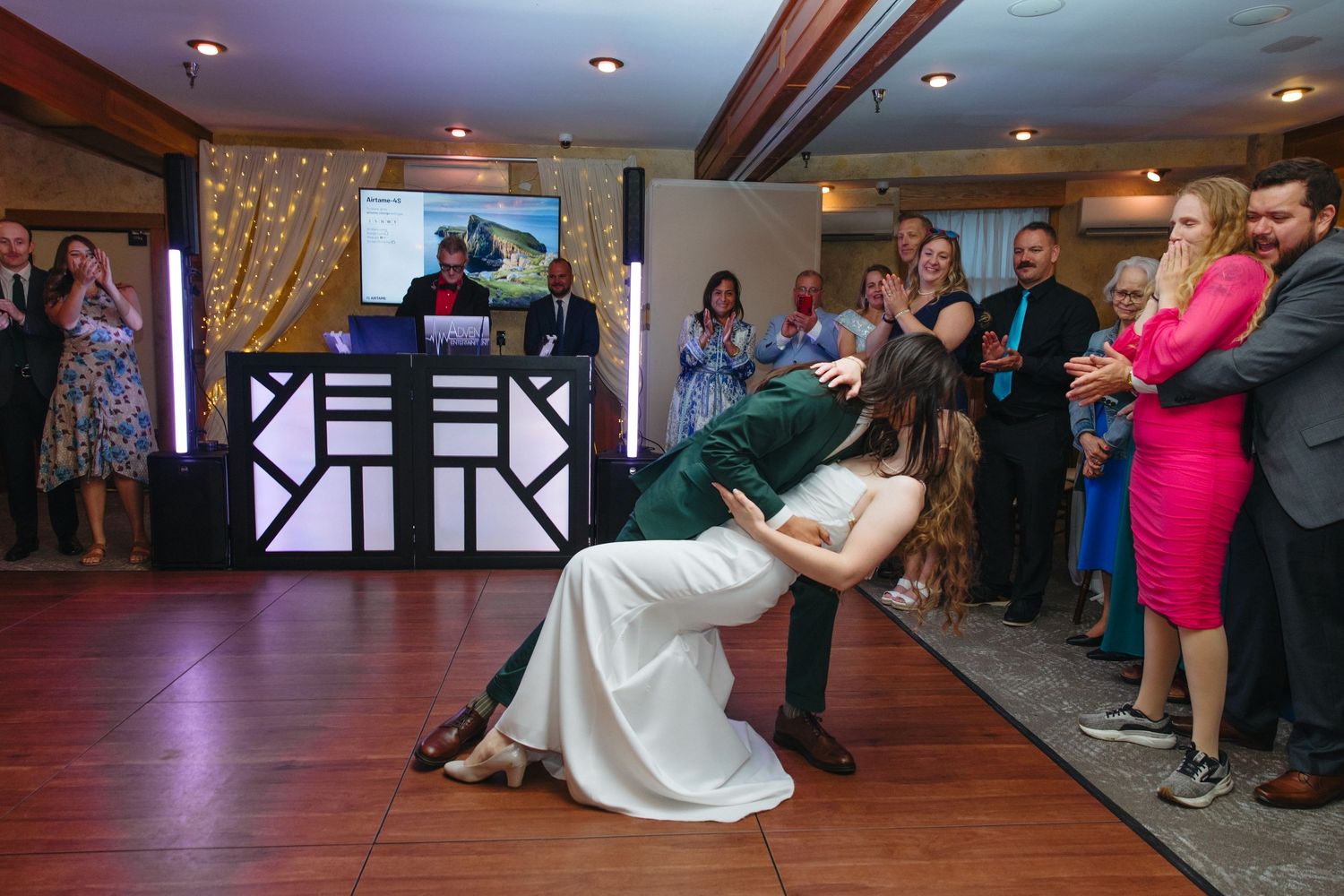 Groom dipping bride during first dance at wedding reception with guests watching and cheering.