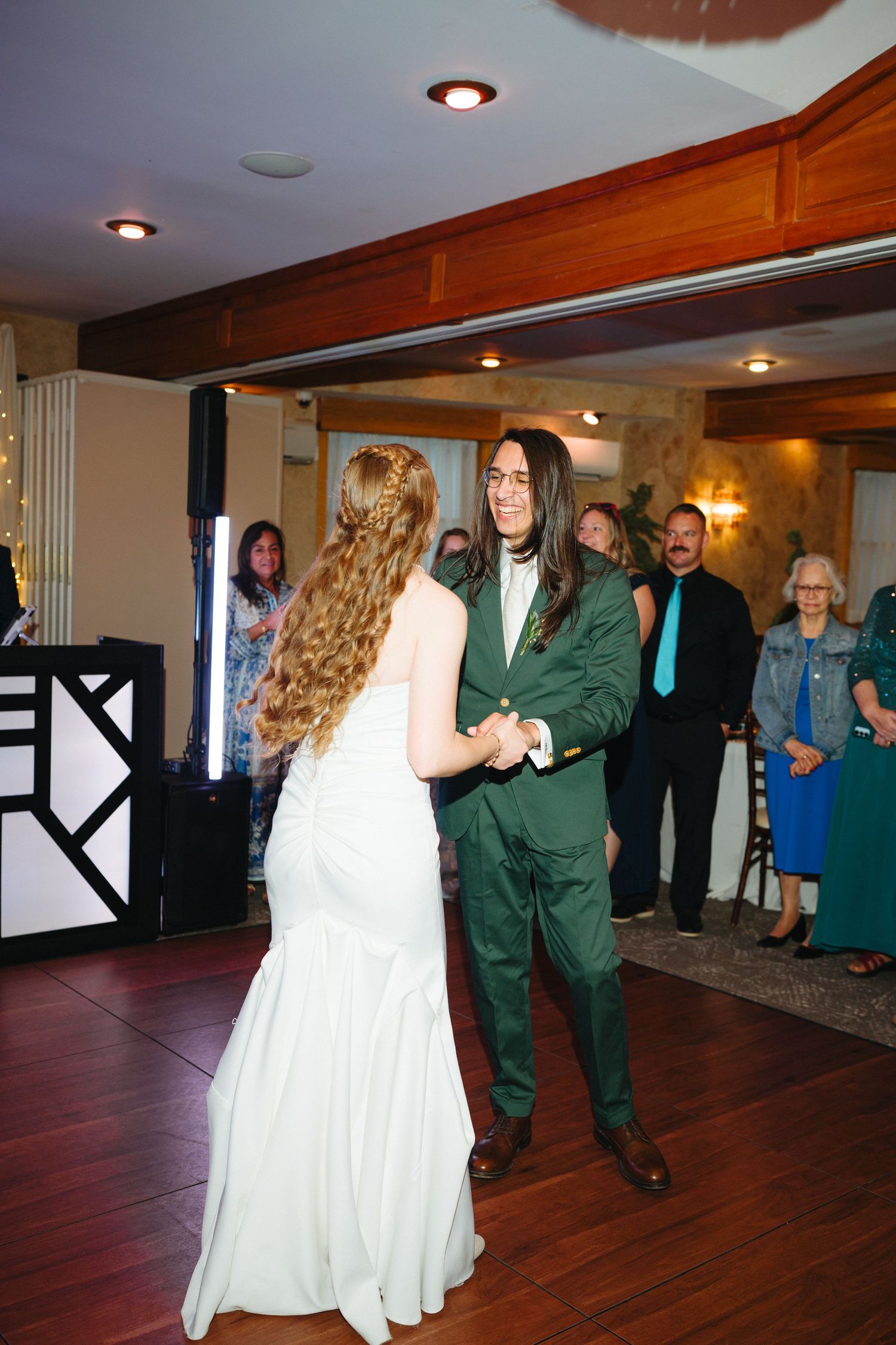 Bride and groom share first dance at wedding reception, groom wearing green suit with long hair.