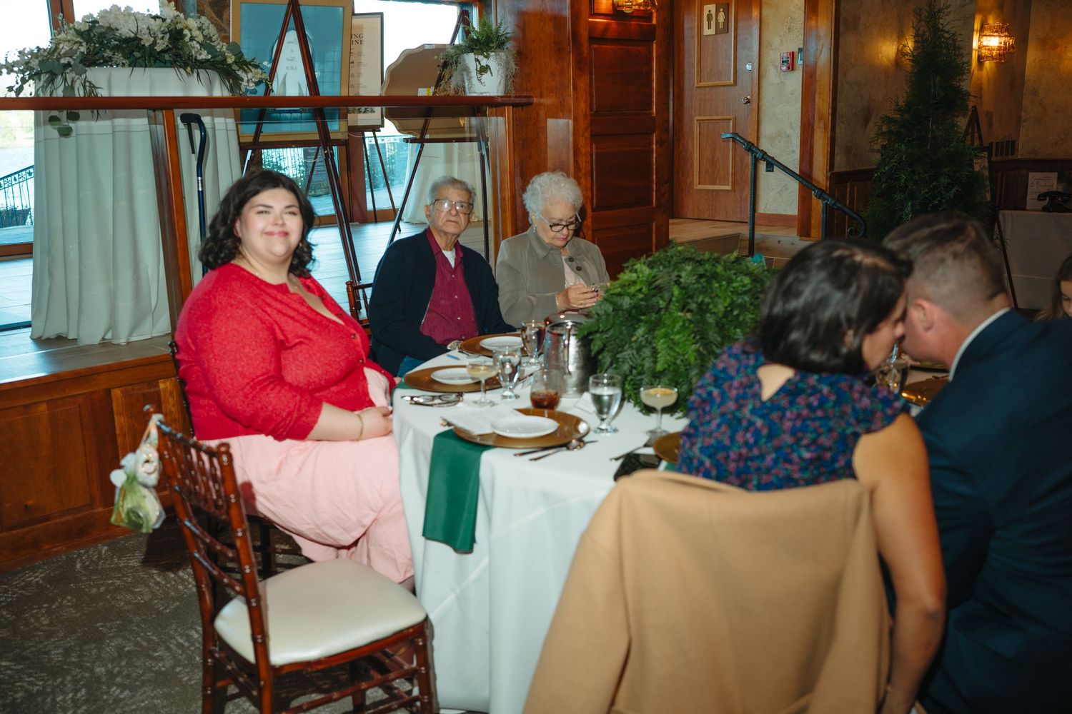 Wedding guests seated at round table with greenery centerpiece during indoor reception dinner.