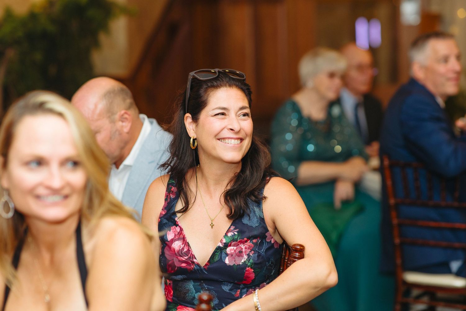 Smiling woman in floral dress enjoying wedding reception with guests in background.