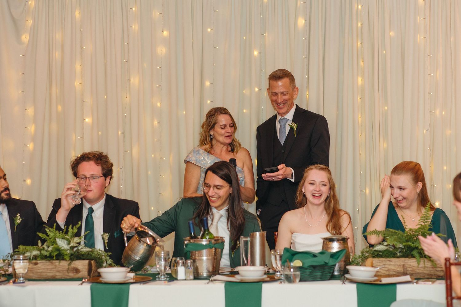 Wedding party laughing at head table during reception speeches with fairy light backdrop.