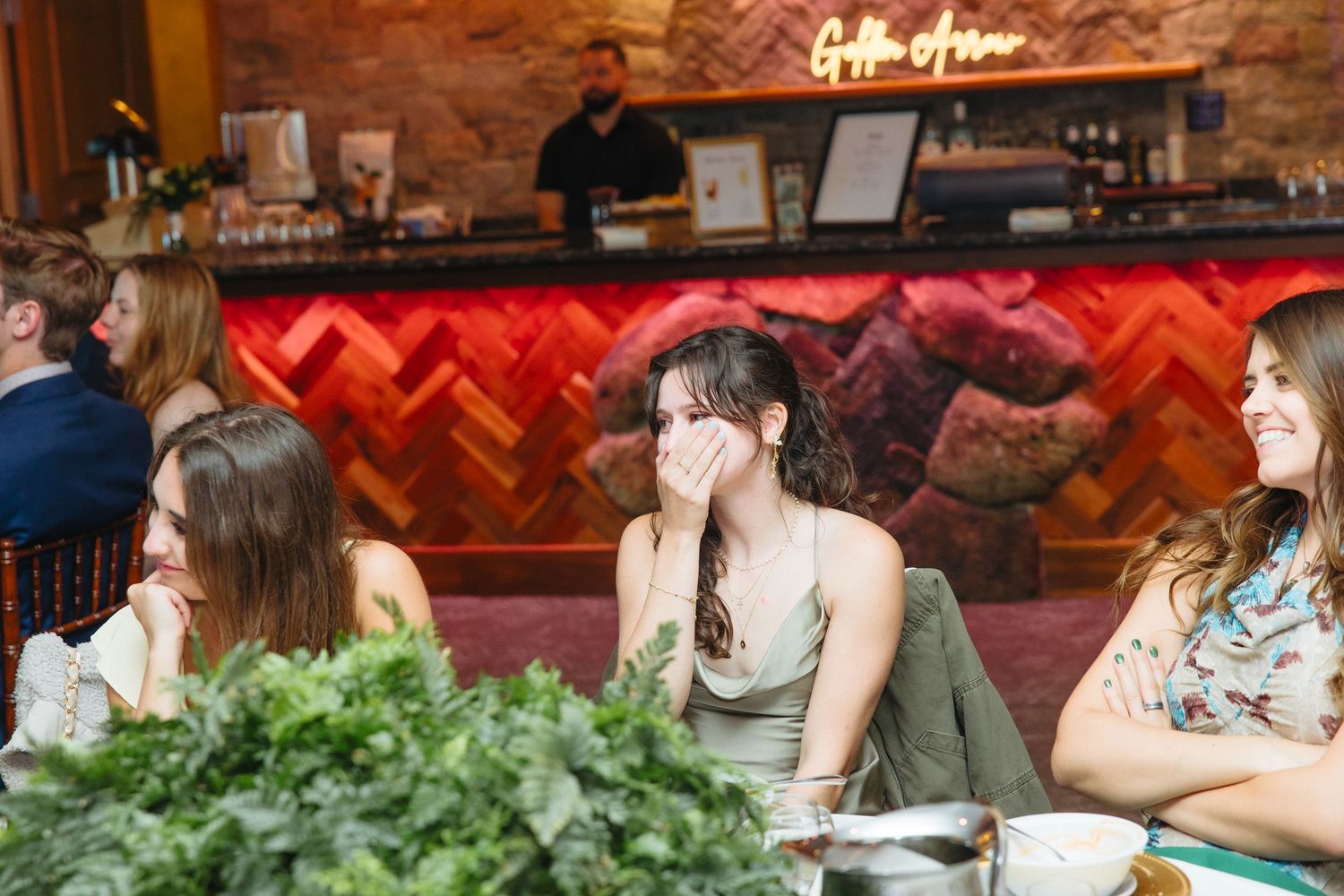 Young women guests laughing and chatting at wedding reception table near colorful bar backdrop.
