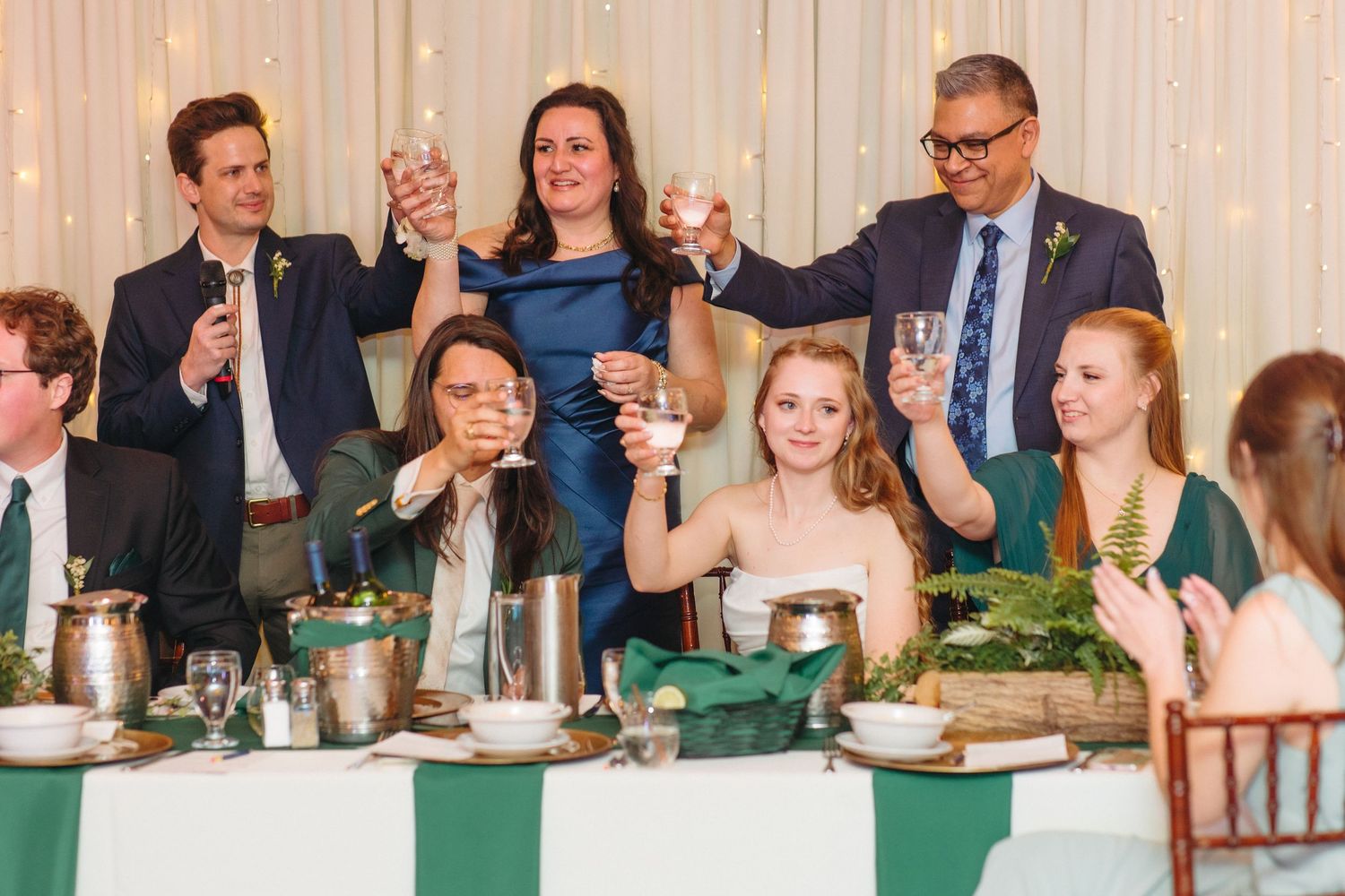Wedding guests raising glasses in a toast at a reception dinner with green napkins and fern decor.