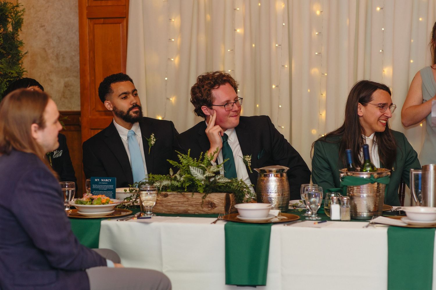 Three guests smile and laugh at a wedding reception dinner table decorated with green linens and fairy lights.