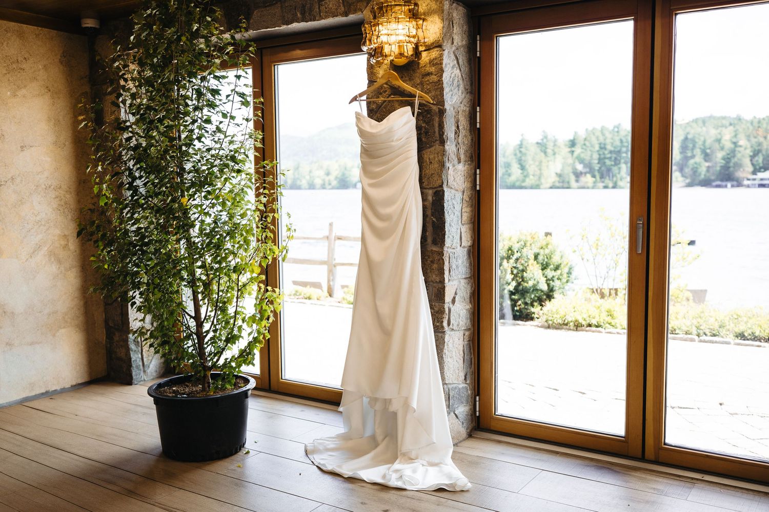 Elegant white wedding dress hanging near large windows with scenic lake and forest views in rustic venue.