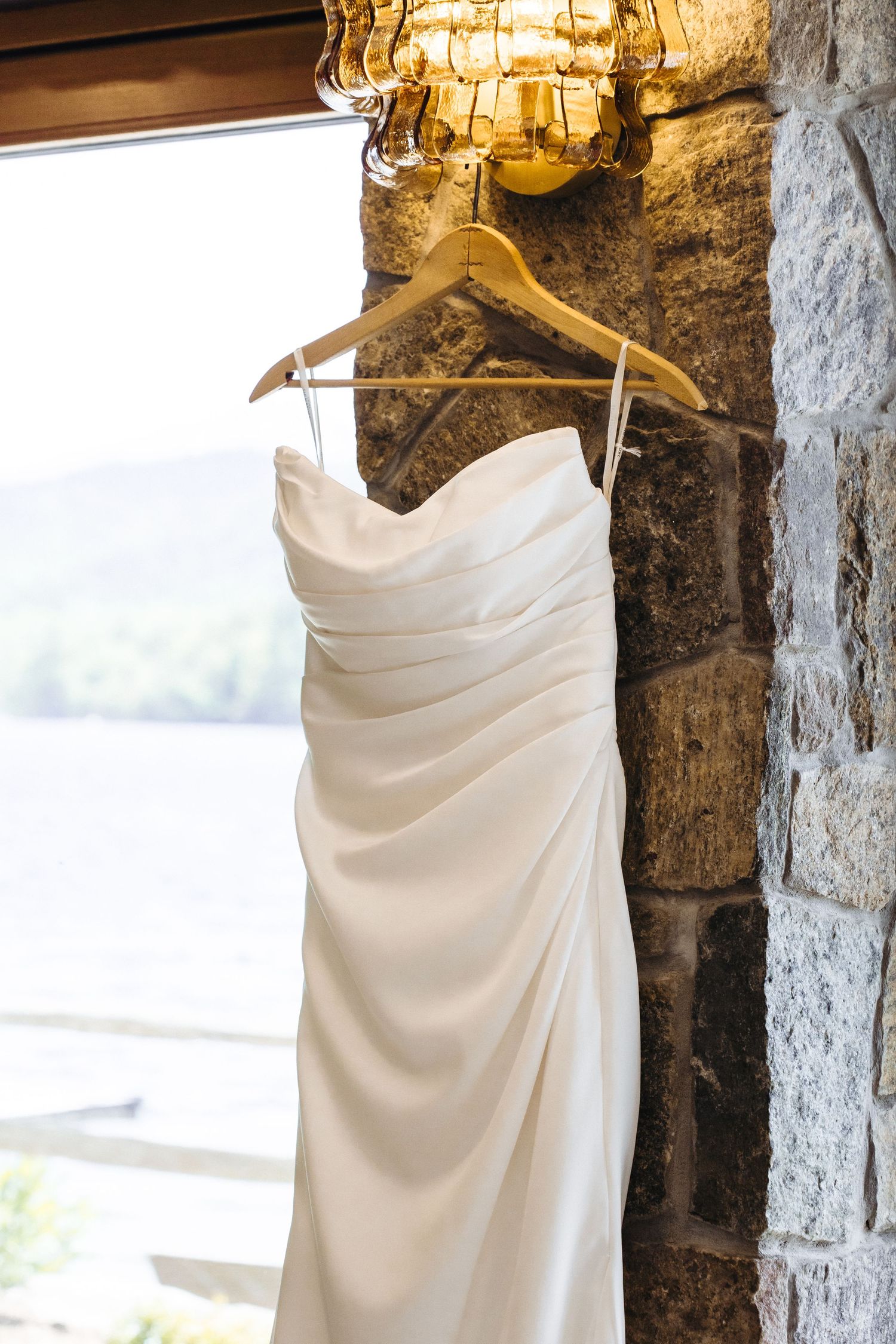 Elegant white strapless wedding dress hanging on wooden hanger against rustic stone wall near window.