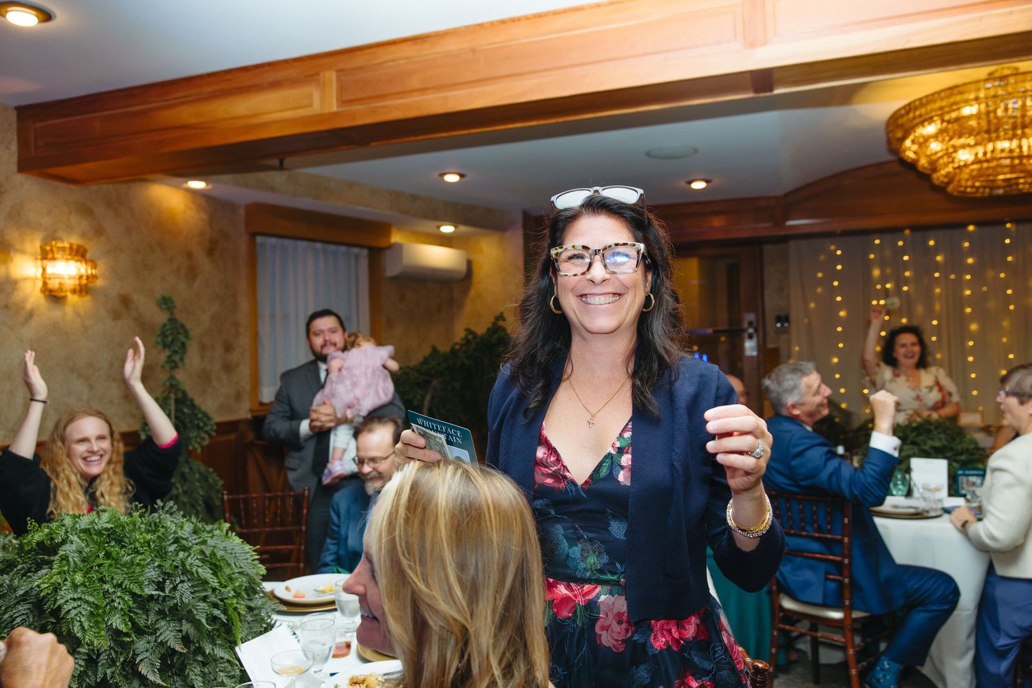 Smiling woman holding small glass celebrating at lively indoor wedding reception with guests.