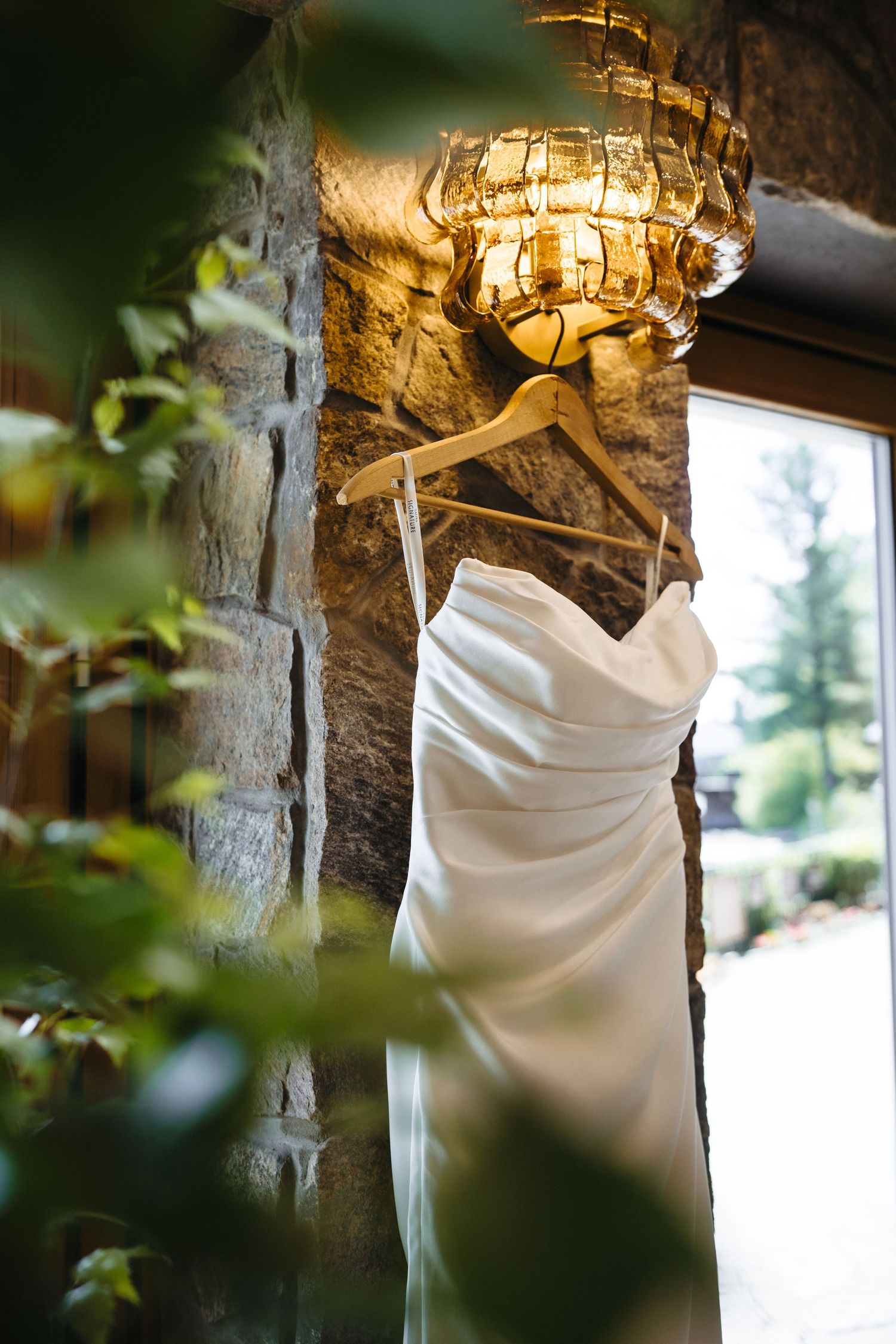Elegant white strapless wedding dress hanging on wooden hanger against rustic stone wall with warm lighting.