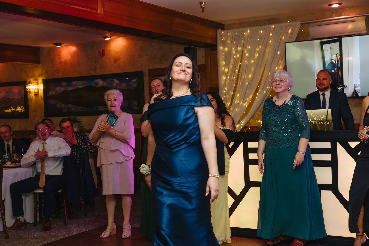 Woman in teal off-shoulder gown dances at wedding reception with guests applauding behind her.