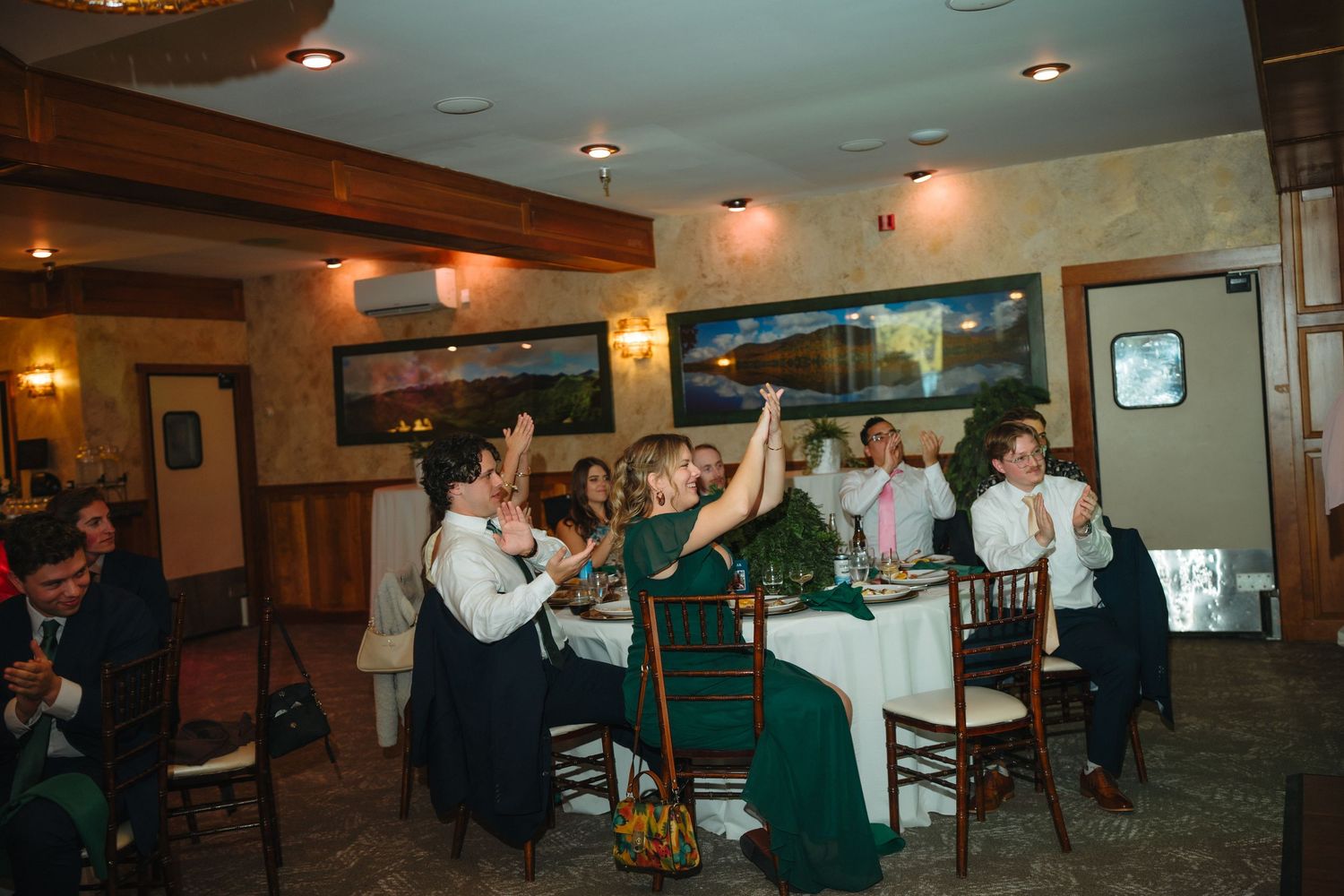 Wedding reception guests celebrate and raise hands at elegantly decorated restaurant venue with warm lighting.