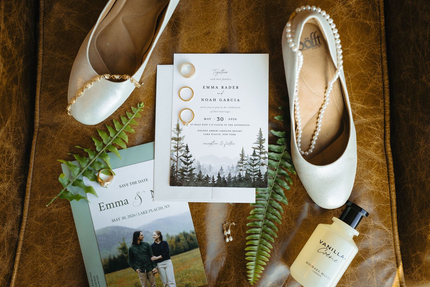Flat lay of wedding invitation suite with white bridal shoes, rings, fern, and perfume on leather surface.