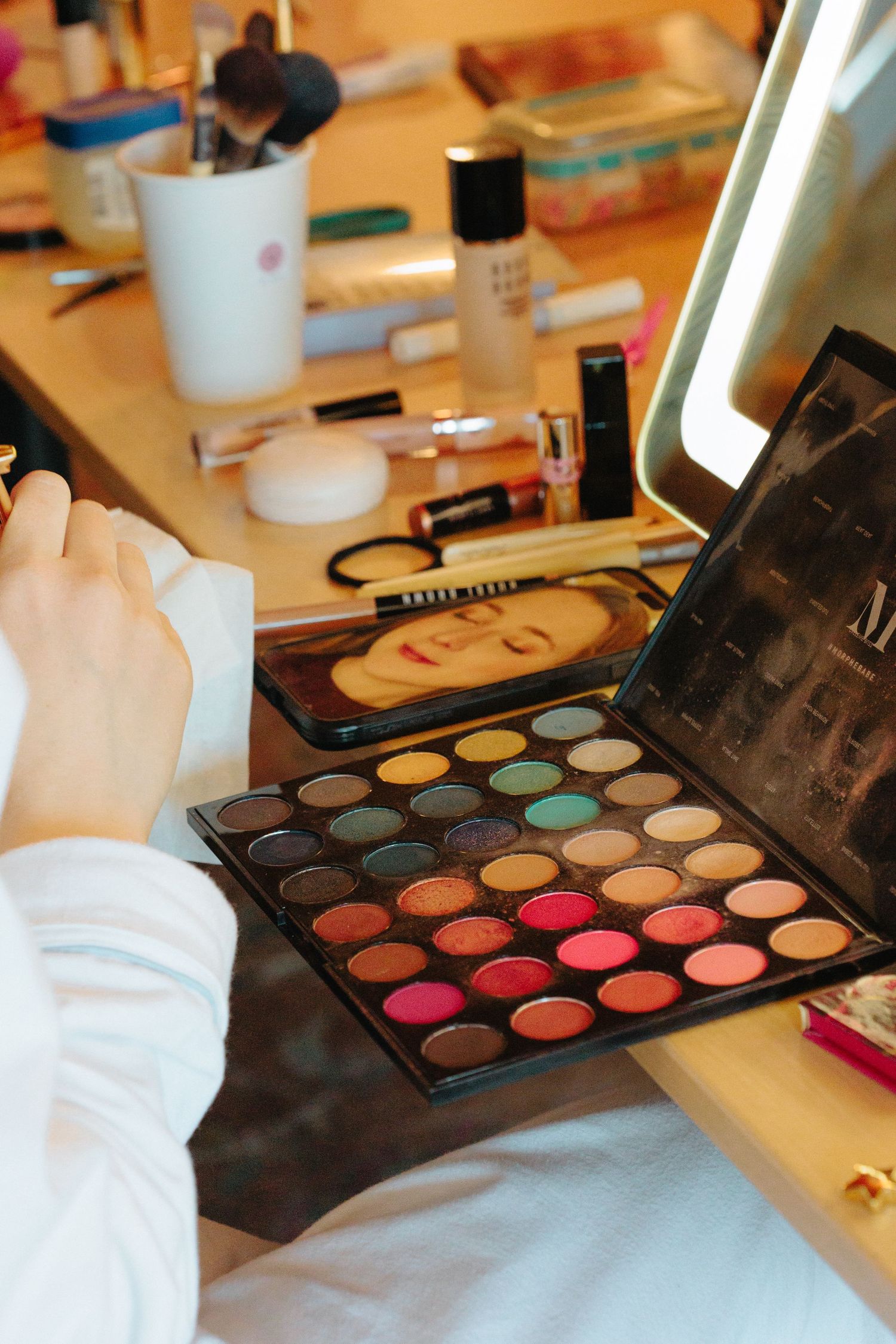 Bride holding a colorful eyeshadow palette at a vanity mirror surrounded by makeup products.