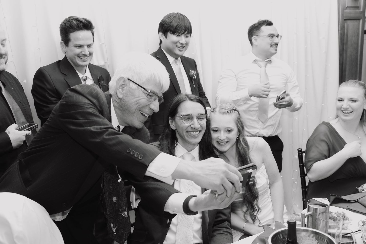 Black and white photo of wedding guests gathered around smiling couple taking a selfie at reception.