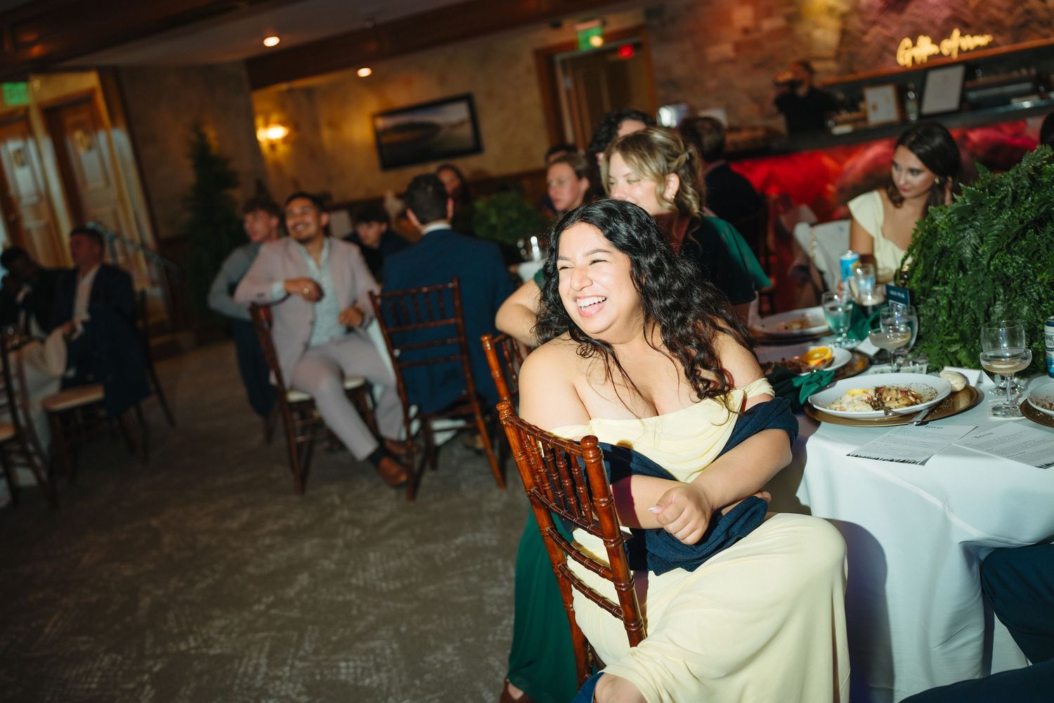 Wedding guest in yellow dress laughs joyfully while seated at decorated reception dinner table.