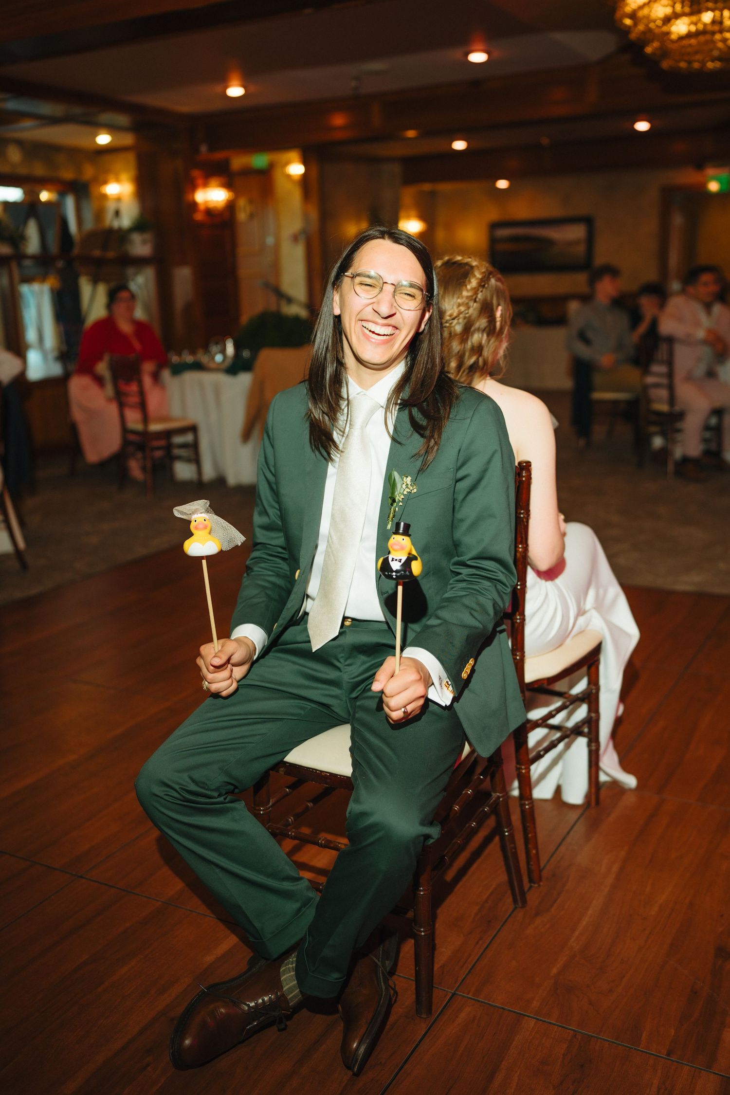 Groom in green suit with long hair and glasses laughing while holding cake toppers at wedding reception.