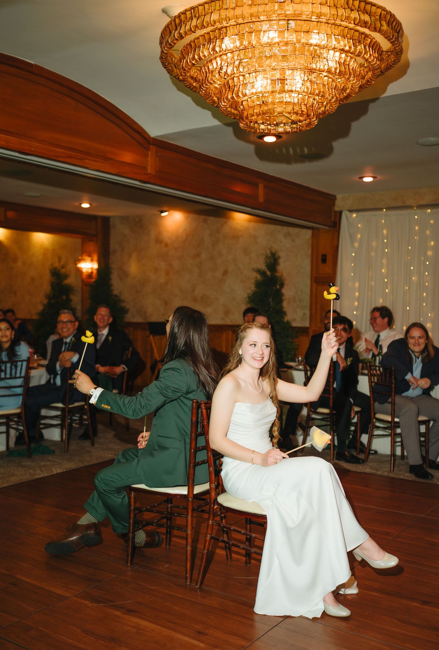 Bride and groom play wedding game seated in chairs under chandelier at reception venue.
