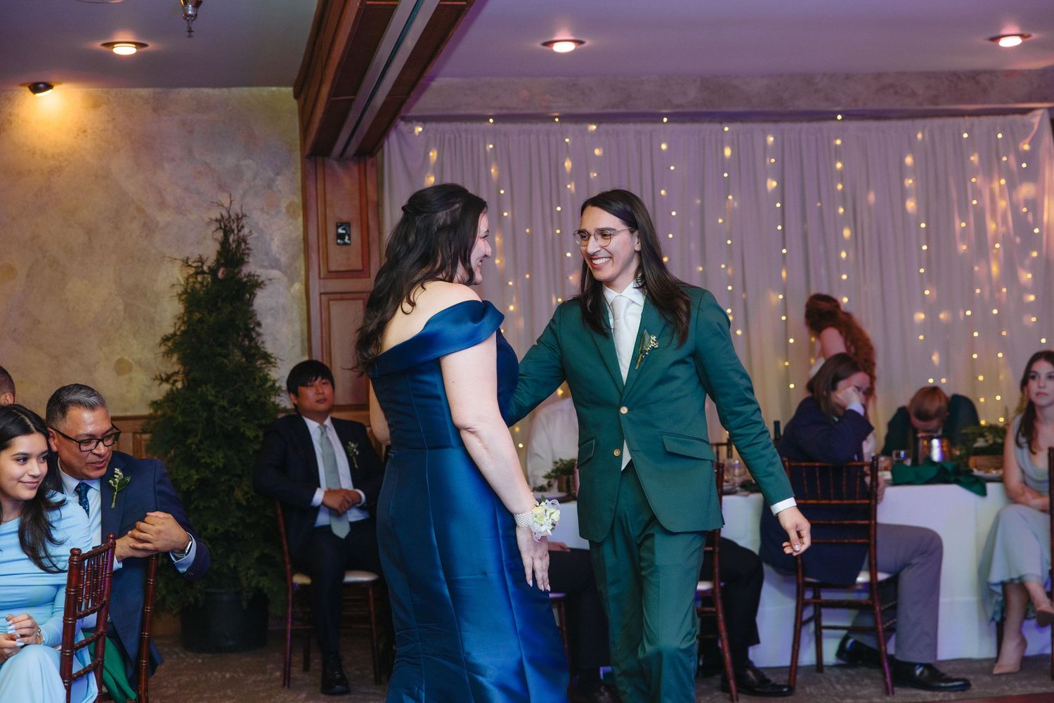 Two women dancing at a wedding reception, one in a blue gown and one in a green suit.