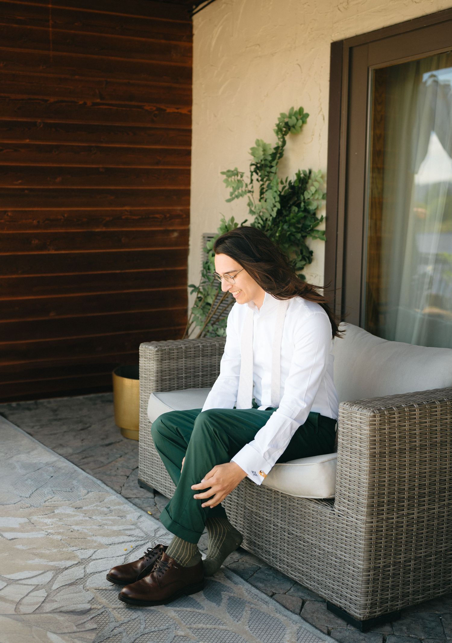 Groom in white shirt and green pants sitting on wicker chair on patio, putting on dress socks.