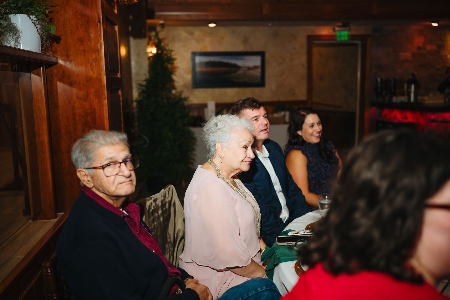 Elderly guests seated indoors at a wedding reception in a warmly lit venue decorated for the holidays.