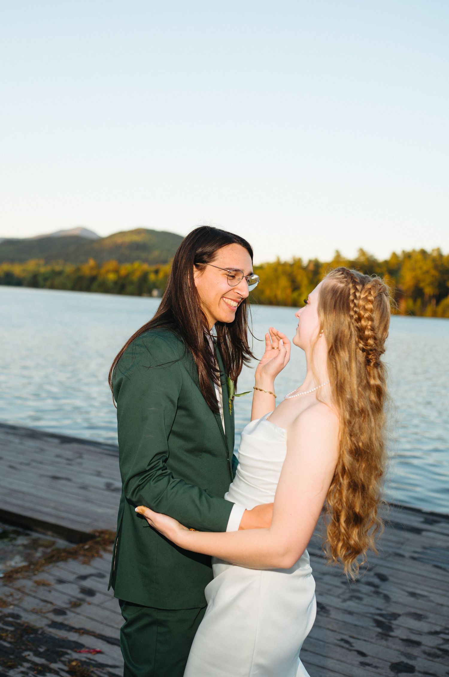 Two brides share a joyful moment on a lakeside dock, one in a green suit and one in a white dress.