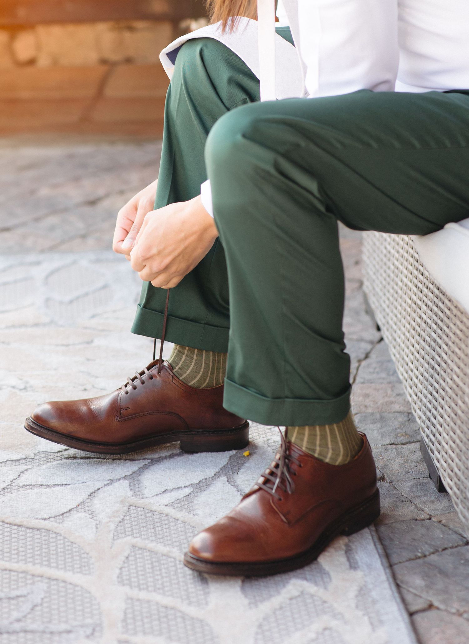 Groom wearing green trousers, striped socks, and brown leather oxford shoes seated outdoors on stone.