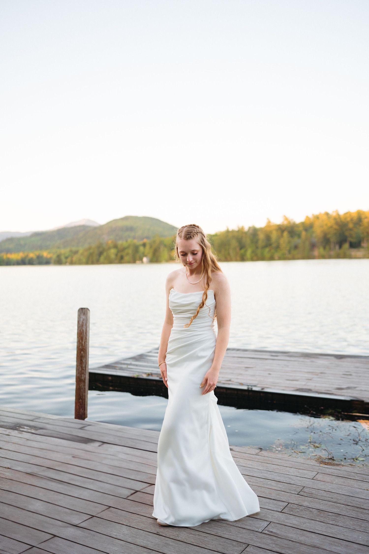 Elegant bride in strapless white mermaid gown stands gracefully on a wooden dock overlooking a serene mountain lake.
