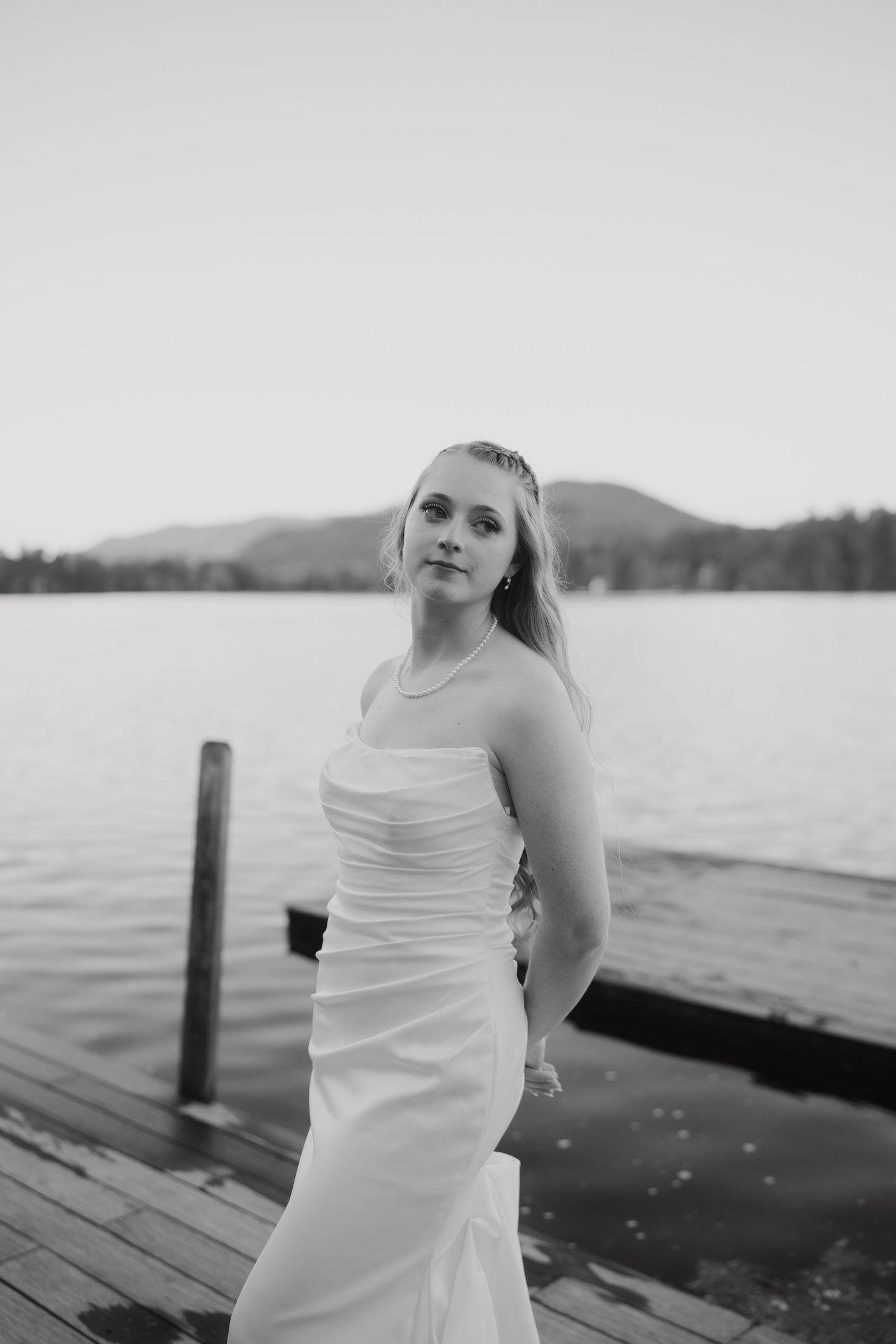 Black and white portrait of a bride in a strapless gown standing on a dock with a lake backdrop.
