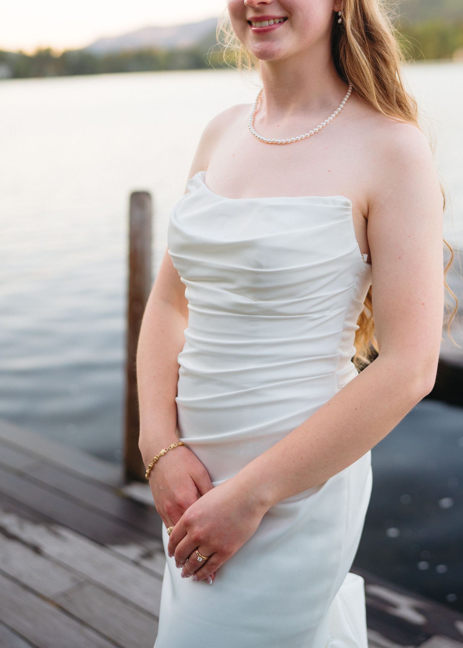Bride in strapless white ruched wedding gown with pearl necklace and bracelet standing on a dock by a lake.