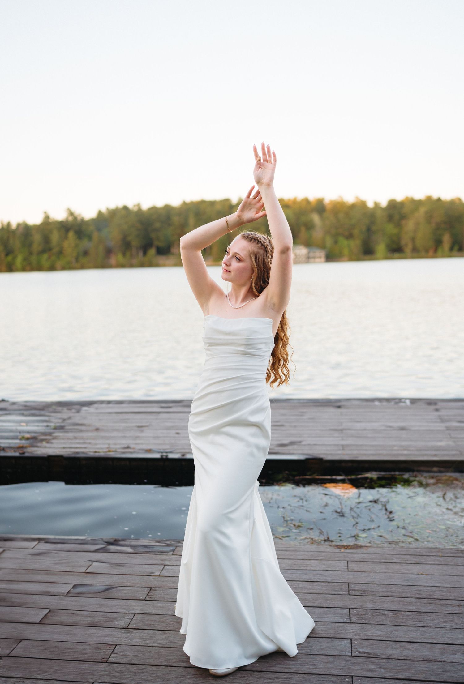 Radiant bride in strapless white mermaid gown poses with arms raised on a lakeside dock at golden hour.