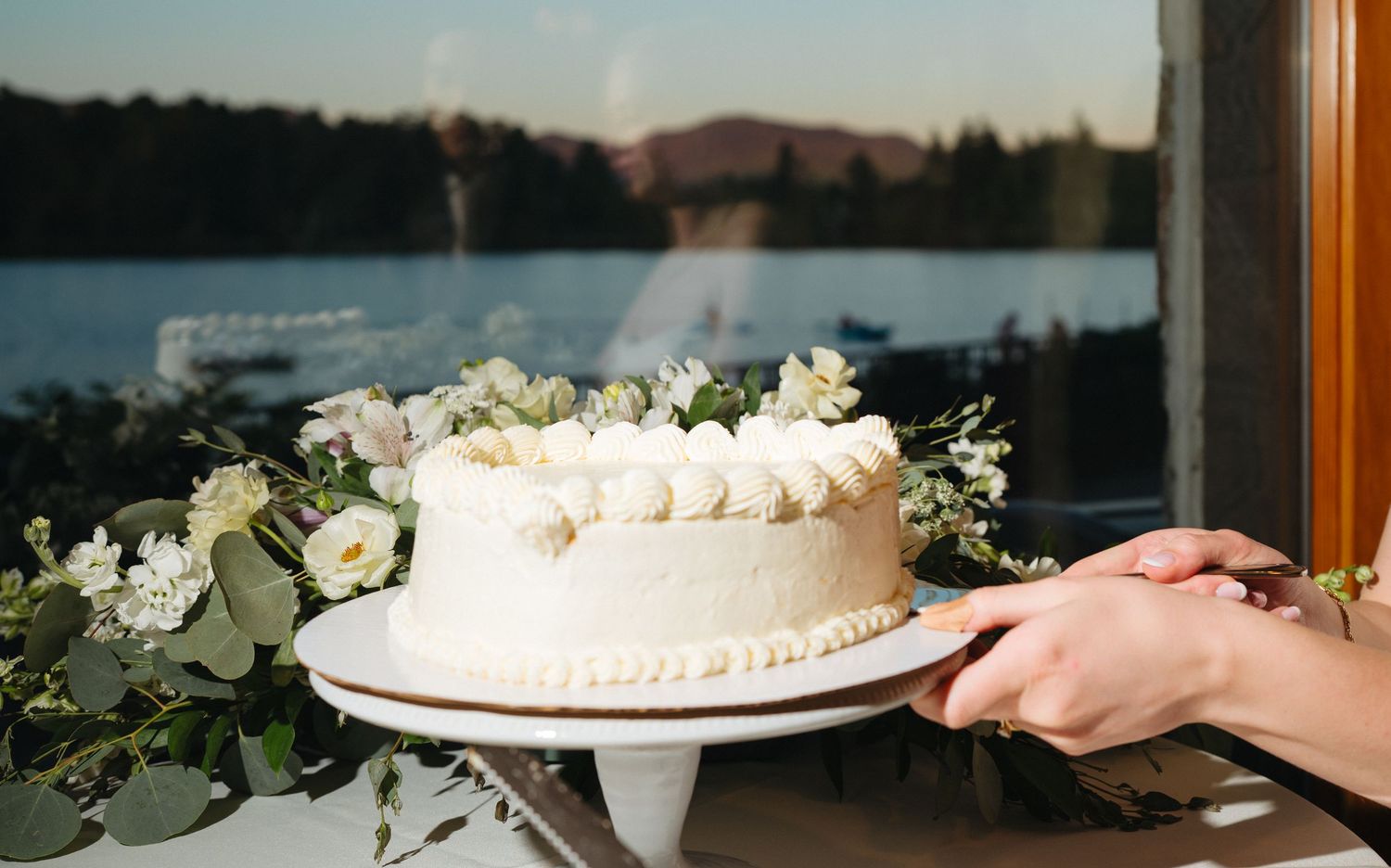 Elegant white wedding cake adorned with fresh flowers on a cake stand, with a scenic lake view in the background.