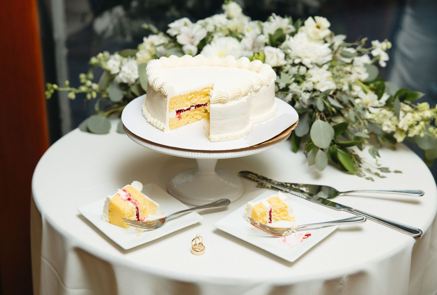 White wedding cake on stand with two sliced pieces on napkins, cake server, and floral arrangement on round table.