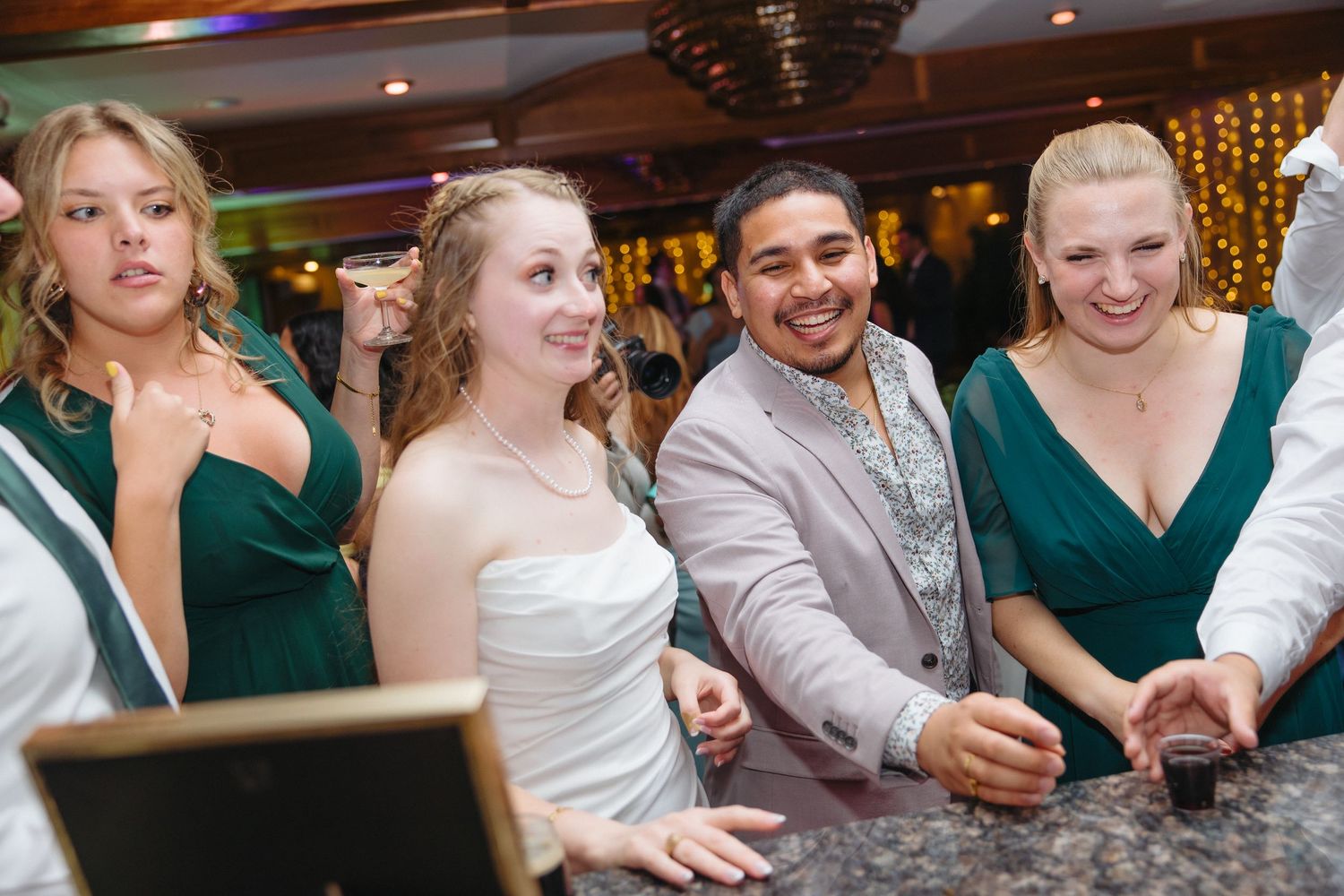 Wedding party smiling at bar, bride in white gown with bridesmaids in emerald green dresses and groom in gray suit.