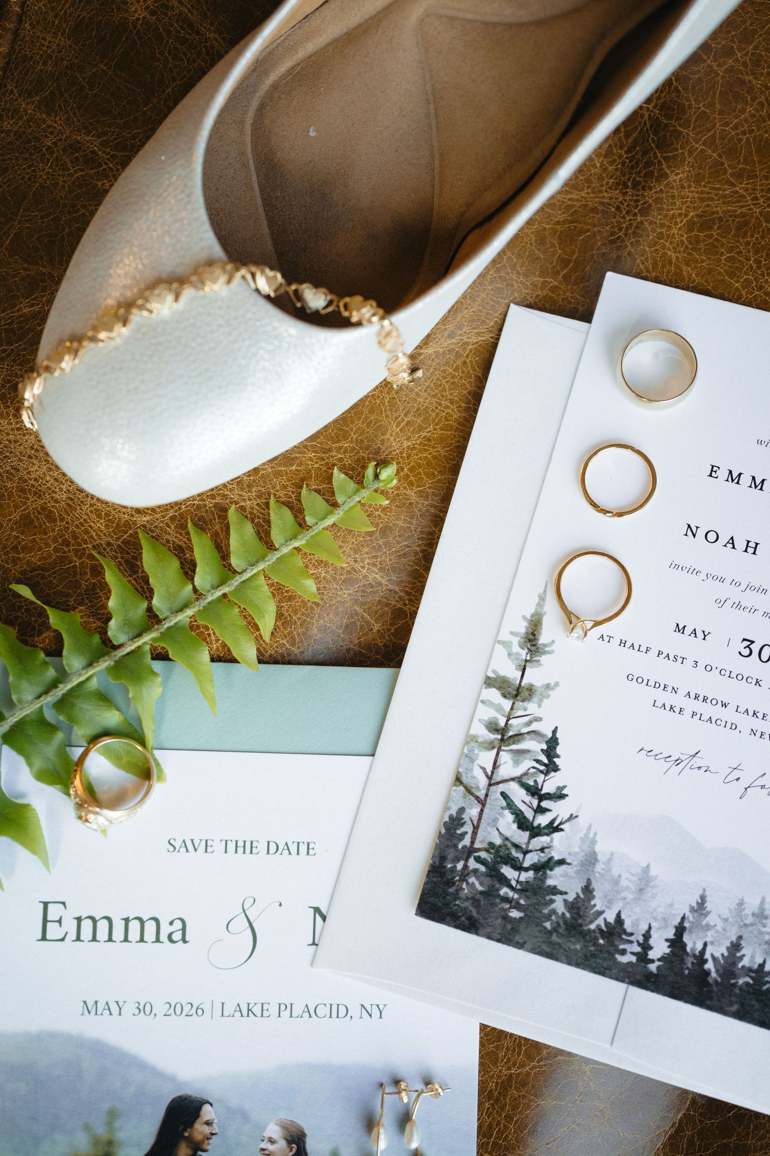 Wedding flat lay with gold rings, bridal shoes, fern, and Emma & Noah Lake Placid NY May 2026 save the date cards.