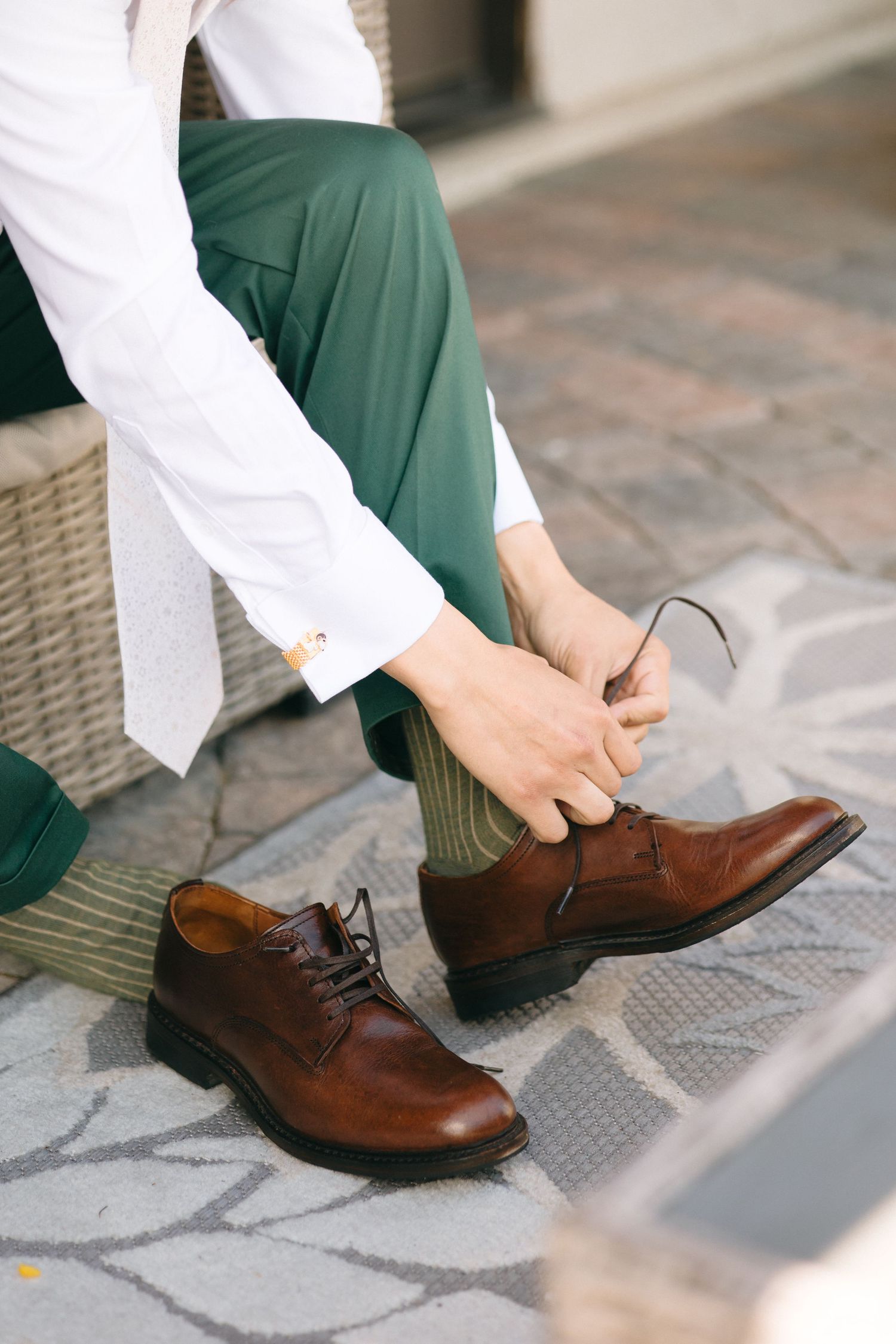 Groom in green trousers and white shirt tying brown leather Oxford dress shoes before wedding ceremony.