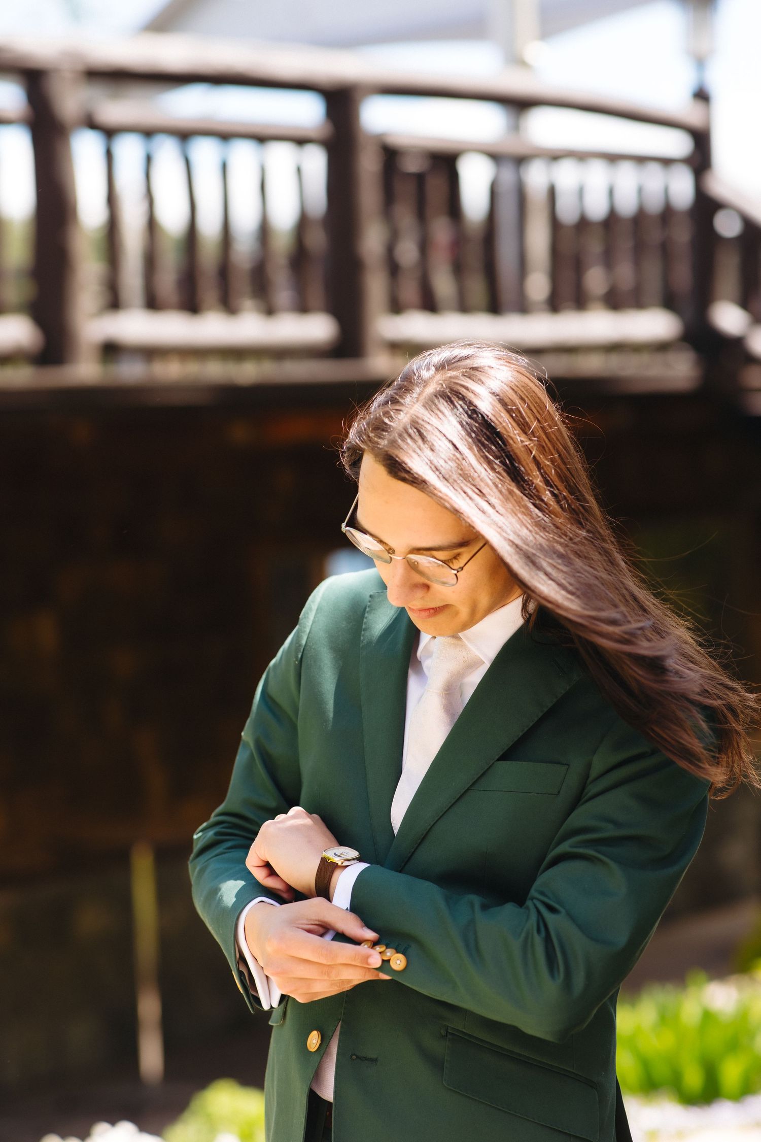 Woman in green blazer checking her wristwatch outdoors near a wooden bridge on a sunny day.