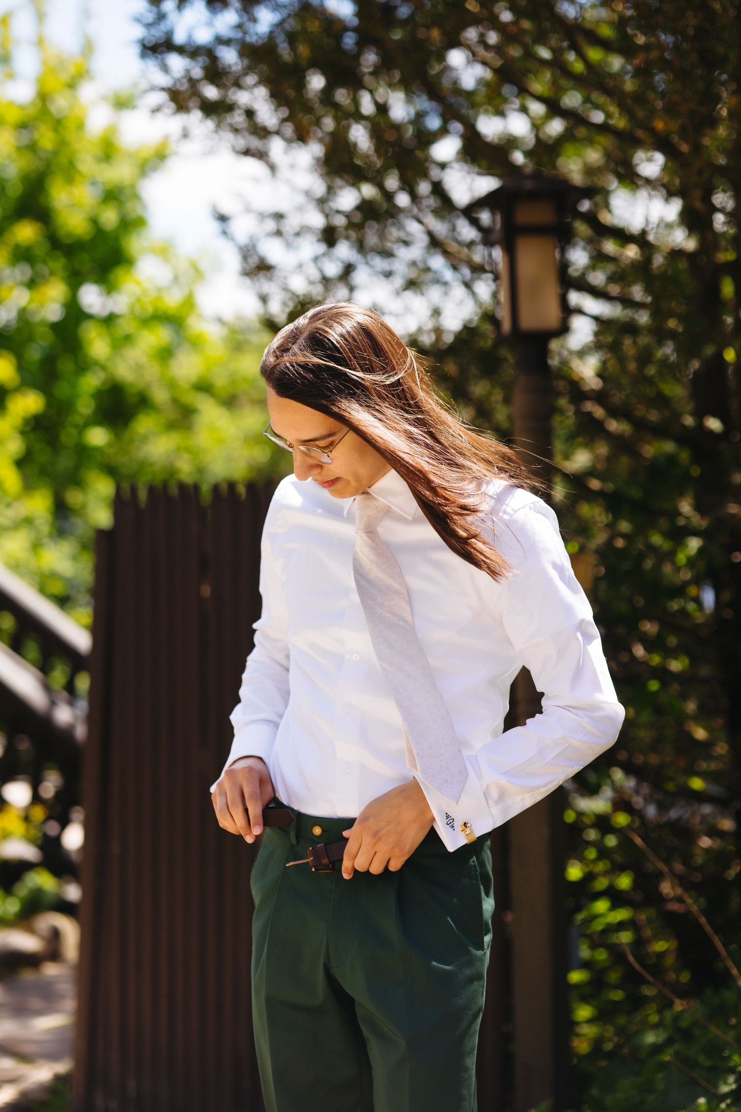 Woman in white shirt and green trousers adjusting her outfit outdoors in a sunlit garden setting.