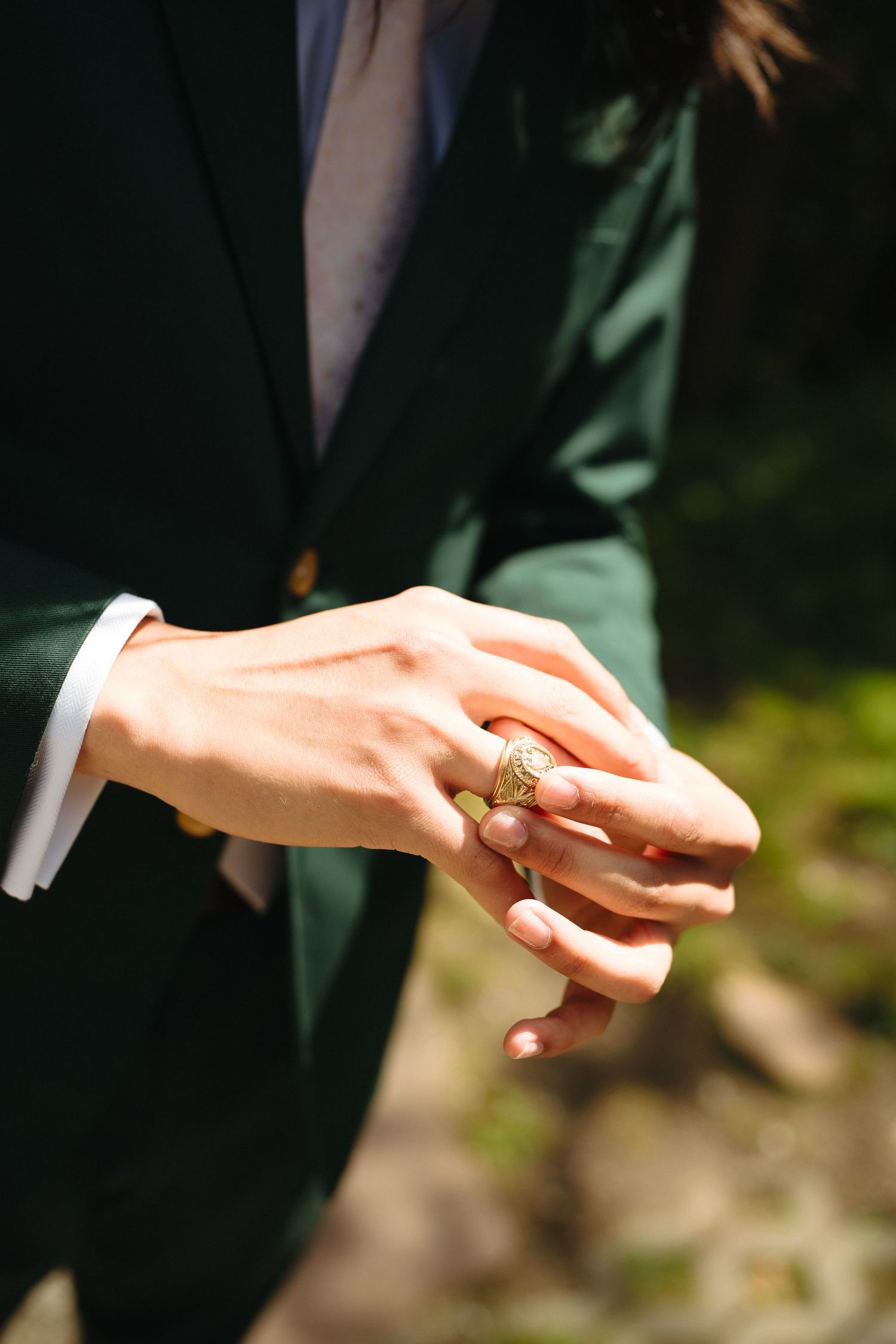 Close-up of groom in dark green suit adjusting ornate gold signet ring on his hand outdoors.