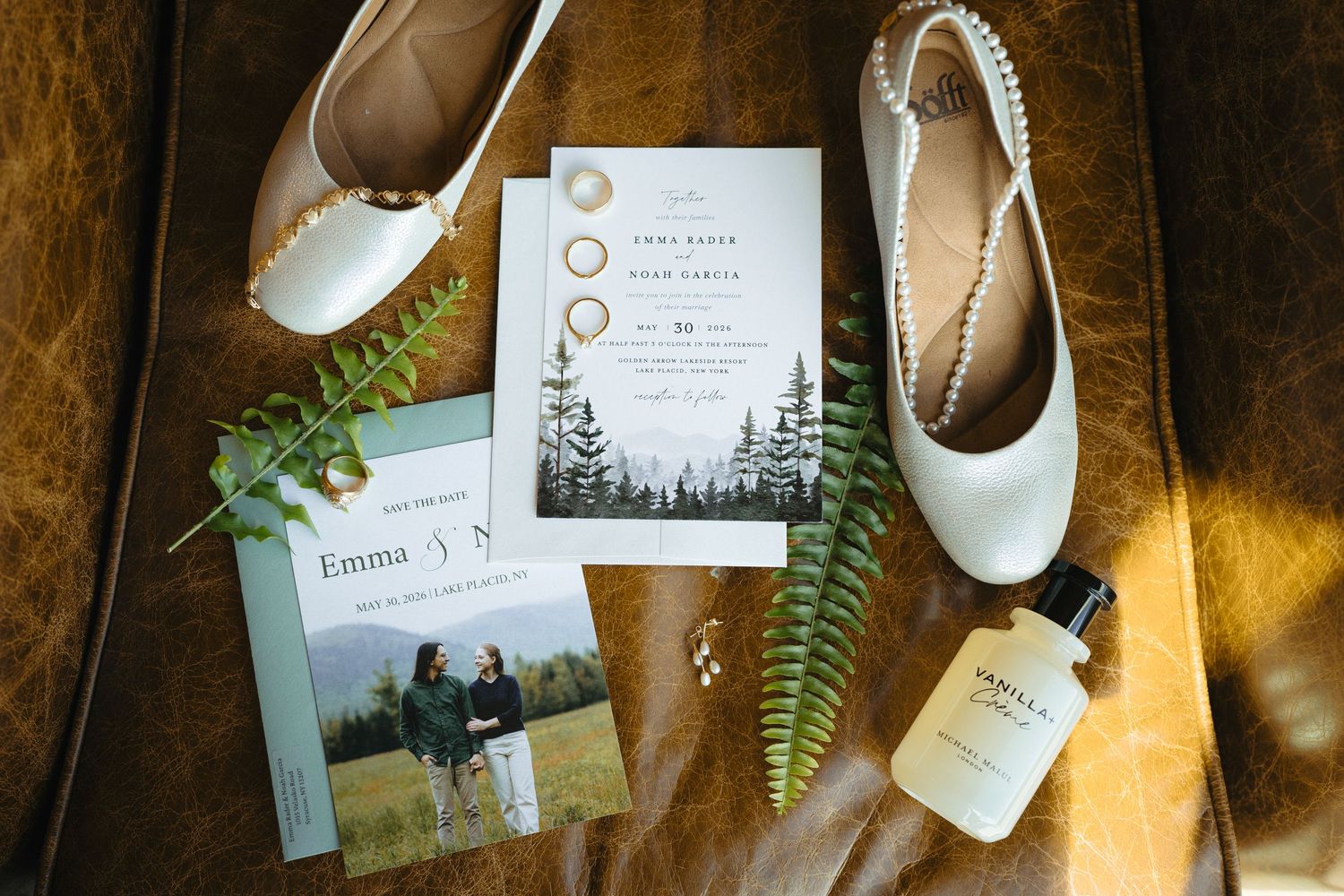 Wedding flat lay with invitation suite, bridal shoes, rings, and greenery on leather surface.