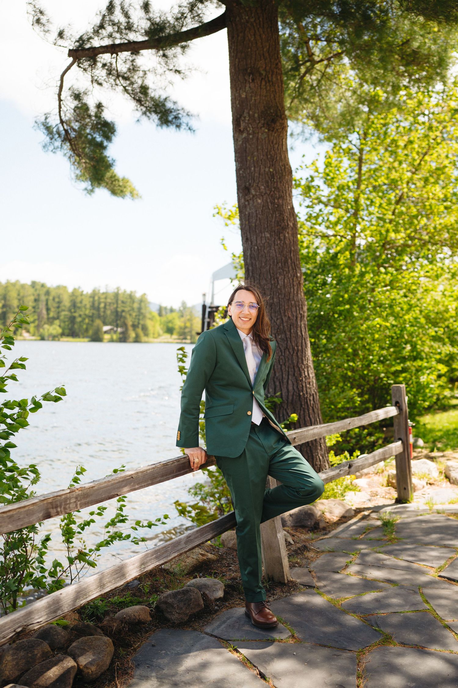 Person in a green suit leaning on a wooden fence by a lake surrounded by lush trees.