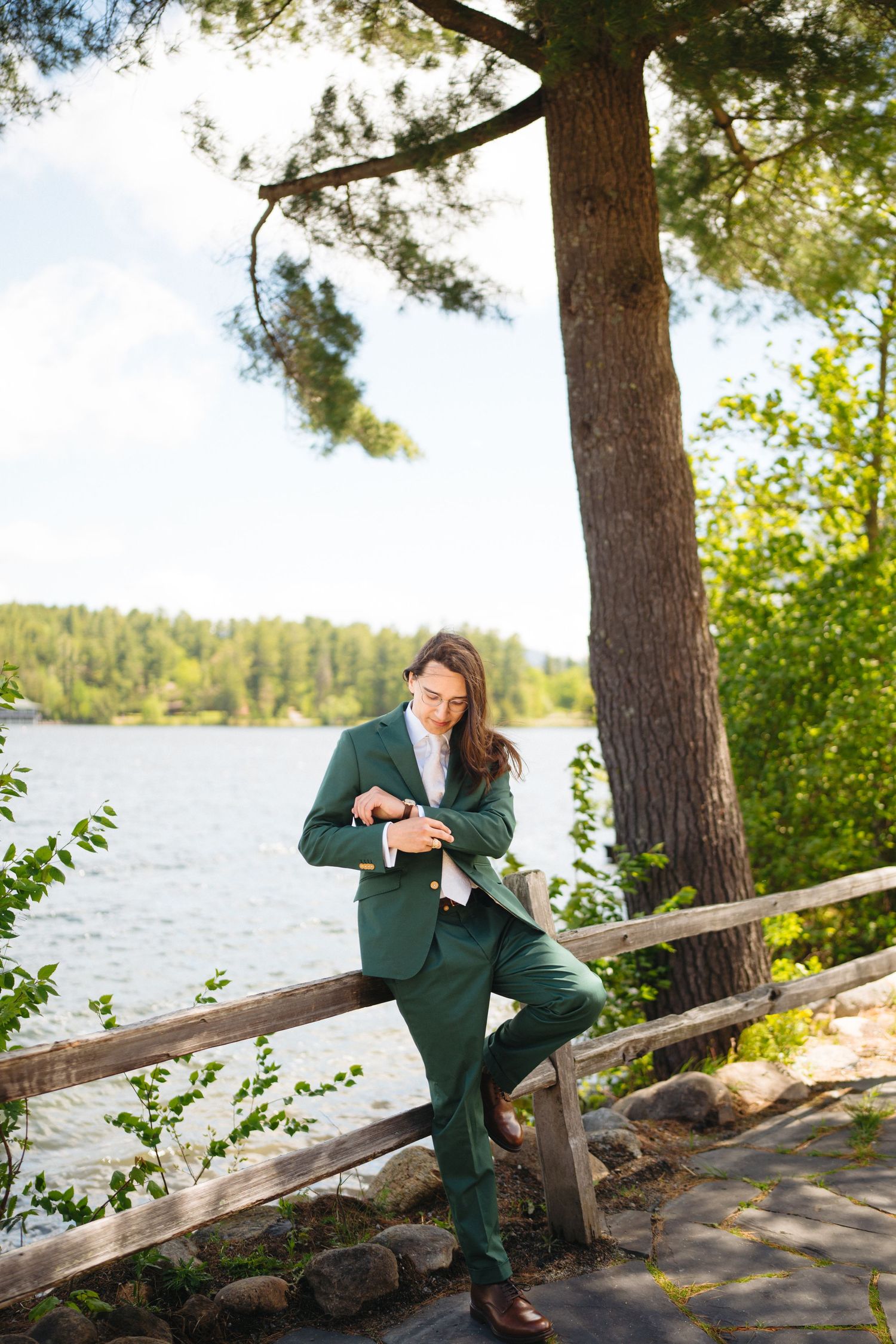 Groom in green suit leaning on wooden railing by a lake surrounded by pine trees on wedding day.