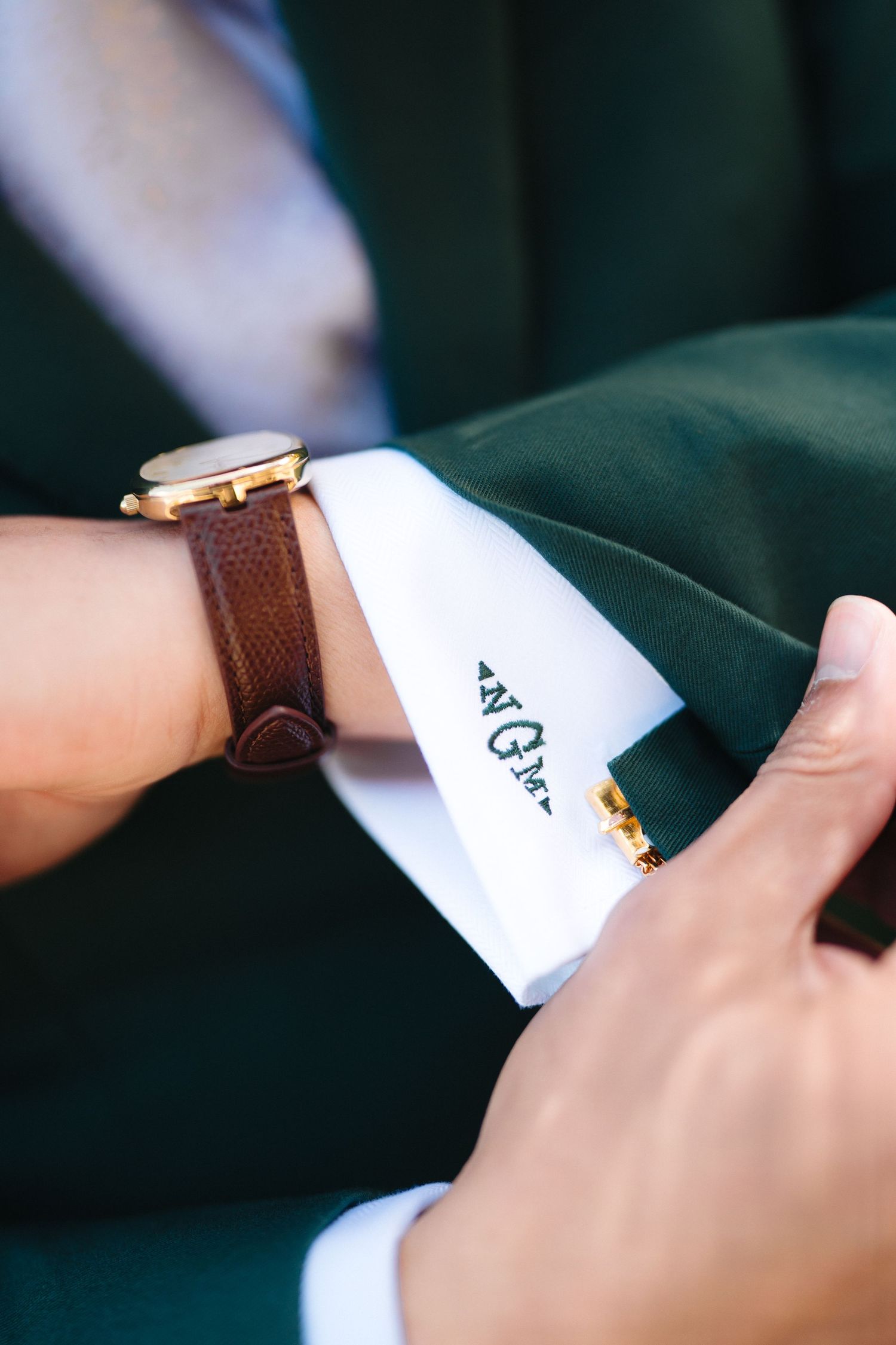 Groom adjusting green suit sleeve revealing embroidered monogram cuff with gold watch and brown strap.