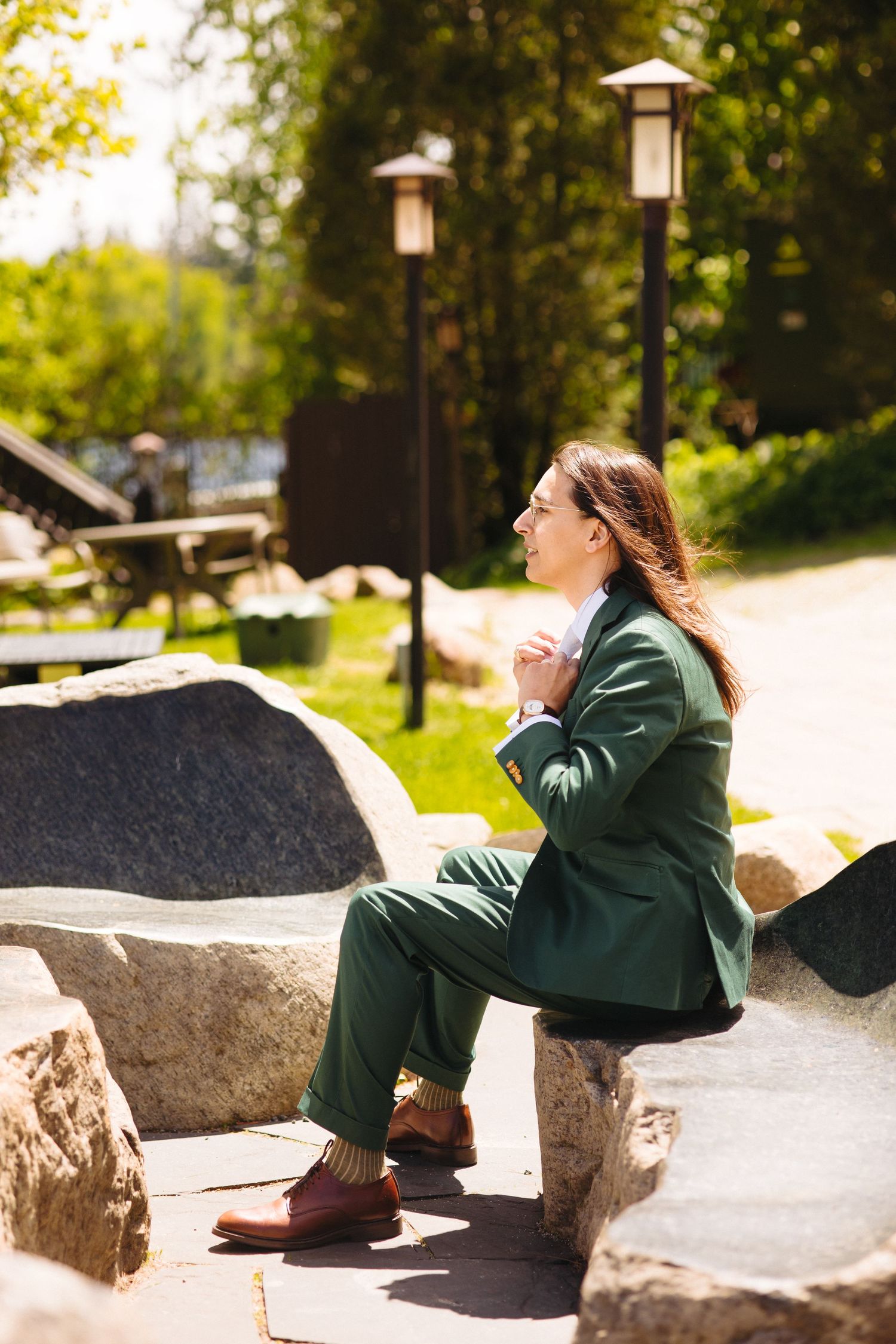 Person in green suit sitting on stone bench in sunny park with lamp posts and greenery in background.