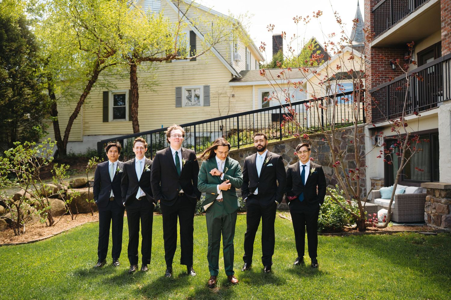 Groom and groomsmen in dark suits pose on green lawn outside a house on a sunny spring wedding day.